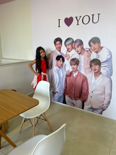 The image depicts an indoor commercial setting in Anápolis, Brazil. A young girl with long dark hair, dressed in a red sleeveless dress with light-colored trim, stands smiling towards the camera. She is positioned to the left of a large, floor-to-ceiling wall decal portraying the seven members of the South Korean K-pop group BTS. Her right hand is placed on the shoulder of one of the decal figures, and her left hand rests on her hip. The decal shows the BTS members in various light-toned, casual-formal outfits. Above the decal, the phrase 'I ❤️ YOU' is printed in black, with a purple heart symbol. In the foreground, a light brown wooden table is partially visible on the left, flanked by two white modern chairs with wooden legs. The floor is tiled with light-colored square tiles, and the walls are a uniform off-white or cream color. A low, light-colored ledge is present along the left wall.