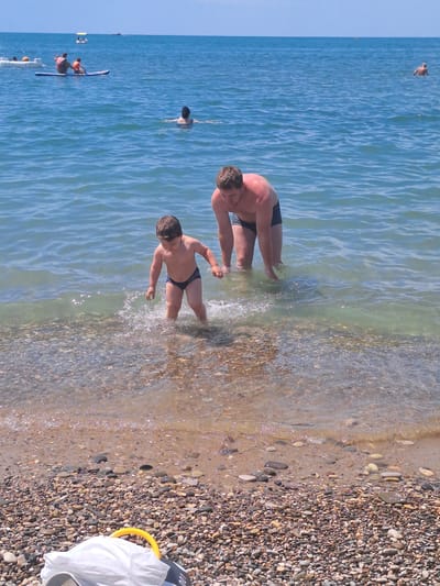 In this vertical, slightly tilted image, a group of people are enjoying a sunny day at the beach in Dzhubga, Russia. The foreground is dominated by a pebble beach, with small, smooth stones of various colors. Gentle waves lap at the shore, creating a shimmering effect on the water's surface.
In the water, a man and a young boy are visible. The man, with his back to the camera, appears to be crouching in the shallow water. The boy, facing towards the camera, is standing in the water, looking down. Both are wearing dark swim trunks. Further out in the ocean, another person is swimming. In the distance, a small group is on a paddleboard or kayak.
The overall scene is one of leisure and recreation on a clear day. The bright sunlight suggests it is daytime, likely in the afternoon given the angle of the light on the water and the intensity of the shadows. The water is a clear, inviting blue, characteristic of a warm, pleasant day. The presence of multiple individuals engaging in water activities indicates a typical summer day at a popular beach destination. There is a yellow ring visible on the shore to the right of the frame, and a white bag, along with a grey shoe. Part of a black object with the number "100" printed on it is also visible near the bag.