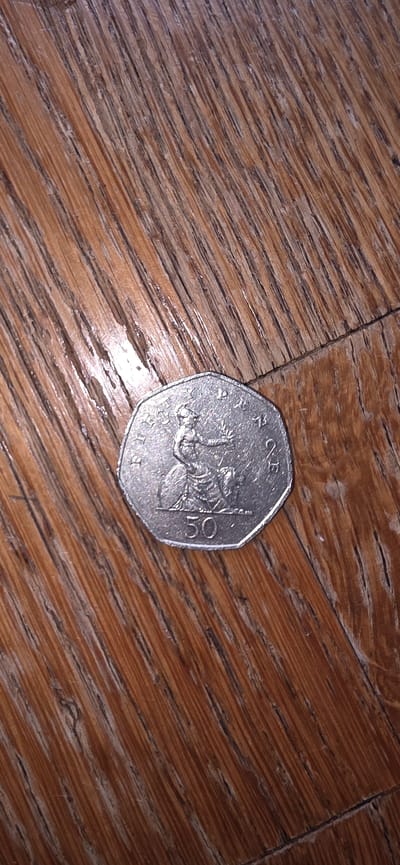 A silver fifty pence coin rests on a wooden surface. The coin features an image of Britannia on its obverse side, a common motif on British currency. The number "50" is clearly visible on the coin, indicating its denomination. The wooden surface exhibits a pattern of parallel lines, suggesting it could be a floor or a tabletop. The lighting in the image casts subtle shadows, highlighting the texture of both the coin and the wood. The scene is static and appears to be a close-up shot of the coin on the wooden background. The context of Marum, Netherlands is not discernible from the visual information in the image.