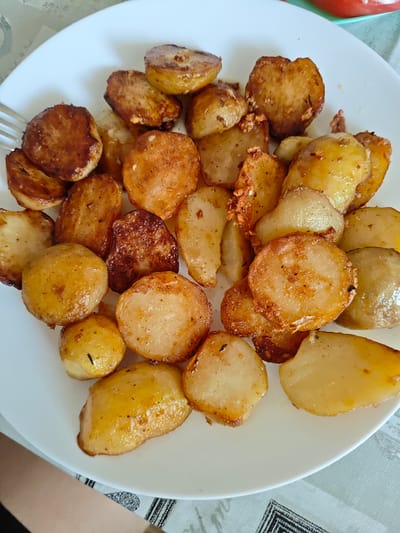 A white plate filled with golden-brown, roasted potato slices is presented in a close-up, overhead shot. The potatoes are seasoned with herbs and spices, and some have slightly crispy edges. A fork is visible on the left side of the plate, suggesting the meal is about to be eaten. The plate sits on a table covered with a patterned tablecloth. The lighting suggests daytime, likely indoors, in a home setting. The potatoes appear to be a main dish or a side, prepared for a meal. The overall impression is of a simple, hearty, and appetizing dish. There is no visible text in the image.