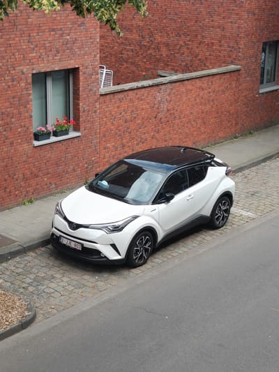 A white Toyota C-HR hybrid vehicle with a black roof is parked on a cobblestone street next to a red brick building. The car has license plate number "2-JJE-803". A blue surgical mask is visible on the dashboard. The building has windows and planters with flowers on the windowsill. The street is paved with cobblestones and a portion of the sidewalk is visible. The image appears to be taken from an elevated perspective, possibly from a window or balcony. The location is the city of Leuven, Belgium.