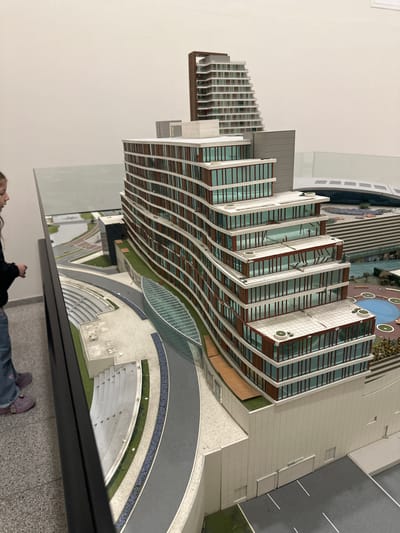 The image shows a scale model of buildings, likely an architectural model. One building is a high-rise with a layered, curving design, featuring numerous windows and balconies. Another building has a terraced appearance. Landscaping elements such as walkways, grassy areas, and perhaps a small pool are visible. A portion of a person wearing jeans and shoes is in the foreground, obscured by a black structure that may be the edge of a table or display case.

Based on the location, the building depicted is likely the architectural model for the Nmerodi Esenyurt in Esenyurt, Turkey.