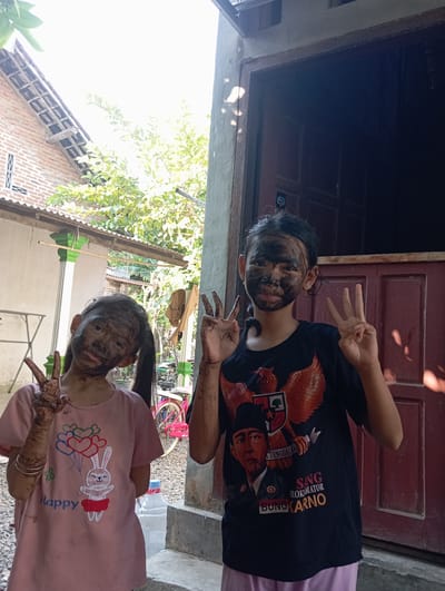 Two young girls, appearing to be Indonesian, stand in front of a rustic wooden doorway in what appears to be a rural or suburban setting. Both girls have their faces and hands covered in a dark, muddy substance, possibly from playing or engaging in an activity outdoors. The girl on the left wears a pink t-shirt with a cartoon rabbit and balloons graphic. The girl on the right wears a black t-shirt featuring a portrait of a man in a hat, an eagle emblem, and text that seems to read "BUNG KARNO." Both girls are making a peace sign with their fingers. In the background, a brick house with a tiled roof is visible, along with lush green foliage and a bright, overcast sky. A red bicycle is partially visible to the right of the girl on the left. The ground is covered in gravel. The overall impression is of a casual, outdoor scene in a warm climate, possibly in the Baron area of Indonesia.