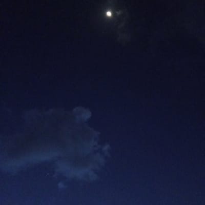 The image shows a night sky with a bright moon near the top center. Wisps of clouds surround the moon. A separate, larger cloud formation is visible in the lower left portion of the frame. A faint point of light, possibly a star, is seen below and to the left of the moon. The sky is a deep blue. Location in Ihururu, Kenya is indeterminable from the image. No people, subjects, objects, actions, or interactions are visible other than the celestial and meteorological phenomena.