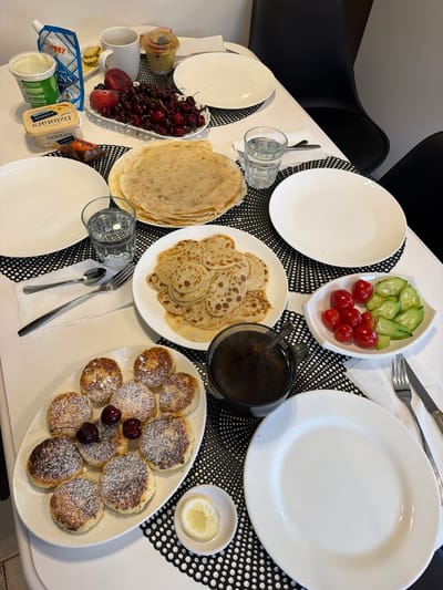 The image shows a breakfast table set for a meal. There are plates, cutlery, glasses of water, and various food items. The food includes a stack of large crepes, a plate of smaller pancakes dusted with powdered sugar and garnished with cherries, a bowl of cherries and a peach, a dish of cherry tomatoes and cucumber slices, a tub of yogurt, a package of butter, a small cup of mustard, and a cup of tea or coffee. The table is set on a white surface with black and white placemats. Two black chairs are visible in the background. The scene appears to be indoors, possibly in a dining area. No people are visible in the image. The location is identified as Baloži, Latvia.