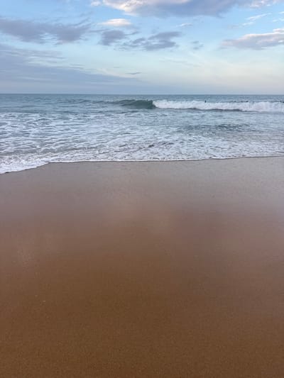 The image depicts a serene beach scene at Arugam Bay, Sri Lanka. The foreground is dominated by the wet, brown sand of the beach, reflecting the soft light of the sky. In the mid-ground, gentle waves with white foam are breaking and receding onto the shore, creating a textured line where the water meets the sand. The ocean stretches out into the background, appearing a calm blue-green. Further out on the water, several surfers are visible, scattered across the expanse, either waiting for waves or riding them. The horizon is defined by the meeting of the ocean and the sky. The sky itself is a blend of pale blue and white, with scattered, wispy clouds that suggest either early morning or late afternoon light. The overall atmosphere is tranquil and natural, characteristic of a coastal landscape.
