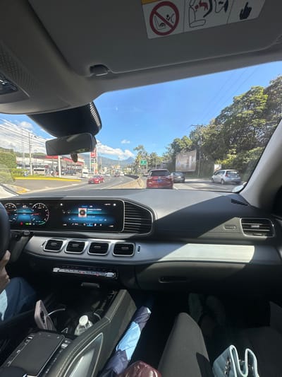 View from inside a car, likely a Mercedes-Benz GLE, showing the dashboard and infotainment system. Outside, a sunny day reveals a street with other cars, trees, buildings, and a gas station visible. Mountains are seen in the distance. The car appears to be at a stop or moving slowly in traffic. An umbrella and a handbag are on the passenger seat.