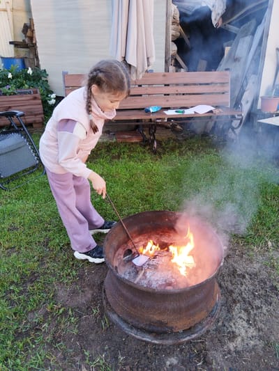 A young girl with braided hair, wearing a pink tracksuit, stands over a rusty metal fire pit in a yard. She is holding a stick with a piece of paper attached, attempting to burn it in the flames. Smoke billows from the fire. Behind her, a wooden bench sits on a grassy area with some plants and a blue barrel. The scene suggests an outdoor activity in a residential area, possibly Sverdlovskiy, Russia, given the context. The ground is a mix of grass and dirt, and there are some wooden structures and a folded canopy in the background.