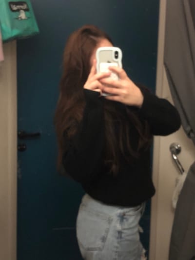 The image depicts a blurry, mirror selfie of a person, presented rotated 90 degrees clockwise. When viewed upright, the image shows a person from the waist up, standing in what appears to be an indoor setting.

The subject is a person with long, flowing dark brown hair that falls over their shoulders and back. They are wearing a long-sleeved black top or sweater and light-wash blue jeans. The person's face is mostly obscured by a white or light-colored smartphone, possibly an iPhone, which they are holding with both hands in front of them, presumably taking the picture in a mirror. The phone has a matching light-colored case, which appears to have a PopSocket-like attachment on the back.

The background elements suggest an indoor environment. Behind the person, there is a dark blue wall or surface. A lighter, beige or off-white horizontal surface is visible below this blue area, which could be a counter, vanity, or floor. In the upper right corner (when upright), a metallic, reflective object, possibly a faucet or part of a light fixture, is visible, along with a greyish surface above it. In the bottom left of the frame (when upright), a light green bag with some indiscernible text or logo is visible. On the lighter horizontal surface in the bottom middle/right (when upright), there are a couple of small, dark-colored objects, possibly bottles or containers.

The image is quite blurry, which, combined with what appears to be low lighting conditions, makes specific details difficult to discern. The primary activity captured is the person taking a mirror selfie. No specific time of day or clear emotions are discernible due to the image quality and framing.