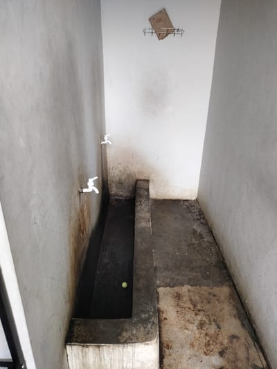 The image captures a narrow, utilitarian indoor space, likely a washroom or laundry area, characterized by unadorned concrete surfaces. The walls are a mix of off-white and grey, showing signs of wear, discoloration, and grime, particularly near the floor and around water sources.
Two white plastic spigots are mounted on the left concrete wall, positioned one above the other. Dark, reddish-brown and black stains trail down the wall beneath these spigots, suggesting past water leakage or frequent splashing. Another similar white spigot is visible on the inner right wall, further back in the scene.
A long, rectangular concrete trough or basin runs along the base of the left wall and extends into the foreground, filled with dark-colored water. A small, light green ball, resembling a tennis ball, floats in the water towards the center of the trough. The edge of the trough forms a low concrete barrier.
On the back wall, a simple metal wire rack is affixed, holding a piece of worn, light brown cardboard. The concrete floor throughout the area is stained and appears dirty, with varying shades of grey and brown. The overall impression is one of a functional but aged and unkempt utility space.
No people are present, and no specific activity is taking place. The scene is static, illuminated by what appears to be natural light from an unseen opening or artificial light. There is no visible text in the image. The location is Dramaga, Indonesia.