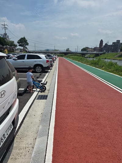 A boy stands next to a small blue bicycle on a paved surface in a parking lot. He is wearing a light blue t-shirt and grey shorts. The parking lot is lined with parked cars. To the right of the parking lot is a red paved path, followed by a green paved path with white stripes. Beyond the green path is a grassy area with a river. In the background, there are buildings, a bridge, and power lines. The sky is cloudy. The location is Anseong-si, South Korea, near the Anseongcheon river.