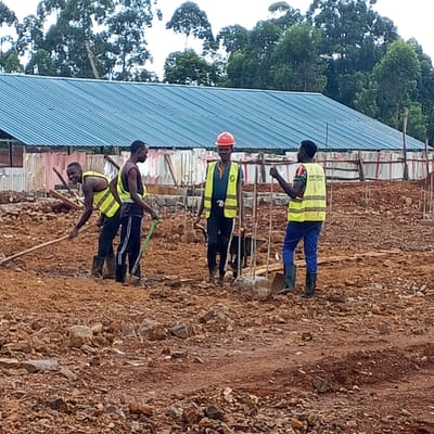 The image shows four men working on a construction site in what appears to be Borabu Chitago ward, Kenya. The ground is rocky and reddish-brown. In the background is a long, low building with a blue corrugated metal roof. The men are wearing safety vests, and one is wearing a red hard hat. They are using tools like shovels and a long wooden pole, appearing to level the ground or prepare a foundation. Vertical metal posts are seen sticking out of the ground. Trees are visible in the background, suggesting the site is on the edge of a developed area.