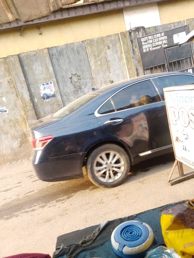 A dark blue sedan is parked on a street alongside a weathered wall with peeling paint and a sign indicating the gate closure times for car owners and general closure. A sign reading "Niferen POS" is propped up on a stand, partially obstructing the view of the car. On the ground in the foreground, various items are scattered on a cloth, including a blue and white object, and a yellow bag. The scene suggests a busy street market or roadside business.