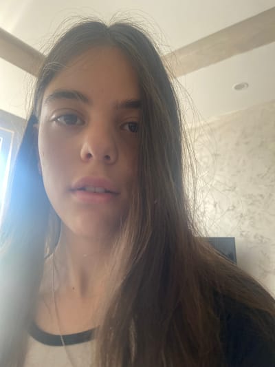 A selfie of a young woman with long brown hair. Her skin appears pale and her eyes are brown. She is wearing a black and white shirt and a thin necklace. The background shows a textured wall and a ceiling with a recessed light fixture. The setting seems to be indoors, potentially inside a home.
There is no indication of location in the city of Dragovishtitsa, Bulgaria.
