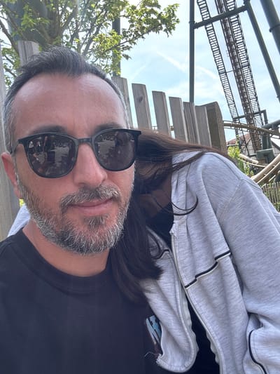 A selfie taken outdoors features a man with graying hair and a gray beard, wearing black sunglasses, a black shirt, and a gray zip-up hoodie. Long, dark hair is visible near his shoulder, suggesting another person is nearby. Reflected in the sunglasses are people and structures, likely indicating a public or tourist area. In the background, there's a complex, geometric metal structure resembling a roller coaster or amusement park ride, along with wooden slats and greenery. The photo appears to be taken in Cleebronn, Germany, based on the presence of the amusement park "Erlebnispark Tripsdrill".