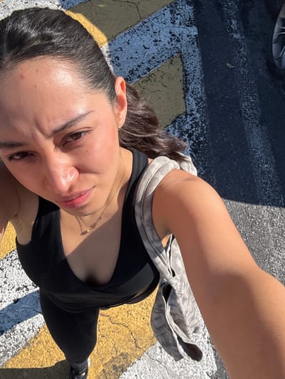 An overhead shot captures a young woman looking up towards the camera, taken outdoors in what appears to be Heroica Puebla de Zaragoza, Mexico. She has dark hair pulled back and is wearing a black sleeveless top or sports bra, and black leggings. A light-colored, possibly grey, patterned garment is draped over her left arm/shoulder. Her expression is a soft smile, and her skin appears dewy from the bright sunlight, with a small red mark visible on her forehead. She wears a delicate gold-colored necklace. The background features dark asphalt with distinct yellow and white painted markings, typical of a street or parking lot. Strong shadows indicate a sunny daytime setting. No visible text is present in the image.