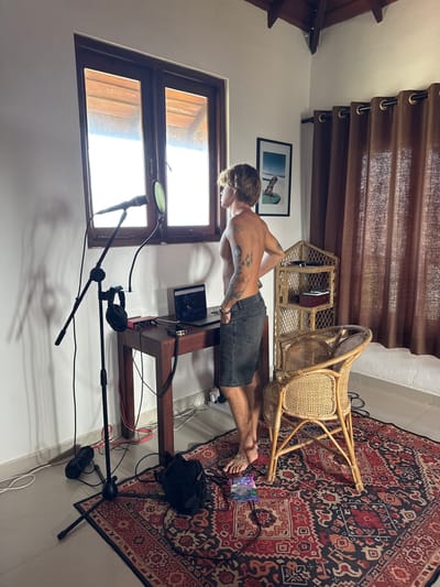 A shirtless man with tattoos on his arms and back stands facing a window, looking out. He is wearing denim shorts. In front of him is a wooden desk with a laptop, a red audio interface, and headphones. To the left of the desk is a microphone on a stand with a pop filter. The room has white walls, a wooden ceiling with exposed beams, and brown curtains covering a window on the right side. A large, patterned rug covers the floor. A wicker chair and a small wicker shelf unit are also present in the room. The scene appears to be a home studio setup. The image is undated and does not contain any explicit identifiers of its location in Goviyapana, Sri Lanka.