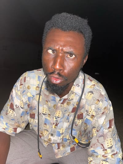 A man stares intensely at the camera with furrowed brows and a slightly open mouth. He has short, dark hair and a well-groomed beard. He wears a patterned button-down shirt featuring building and floral designs. A black cord with yellow accents drapes across his chest. The background is dark, suggesting an outdoor setting with limited lighting. His expression is intense, possibly conveying anger or serious concern.
