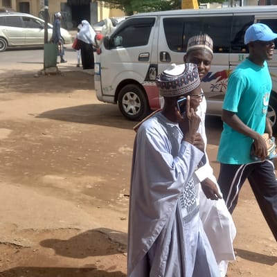 In Dadi Riba, Nigeria, the image shows two men standing outdoors on a partially paved street. One man, wearing a blue traditional cap and a light blue robe, is speaking on a smartphone. The other man, wearing a blue baseball cap and a turquoise shirt, stands nearby. Behind them is a white van with red and black accents. Other people and parked vehicles are visible in the background. The ground is a mix of pavement and dirt.