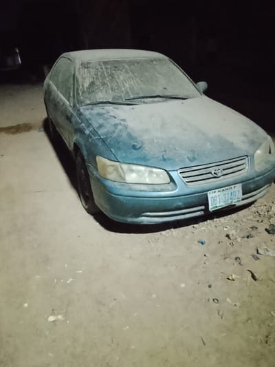 A teal Toyota Camry is heavily coated in dust, obscuring its features. The car's windshield, hood, and roof are almost entirely covered. Its license plate reads "TIO KANO 9 DBT 374BZ." The vehicle sits on a dirt surface with debris scattered around it. It appears to be nighttime.