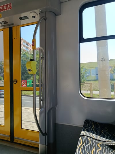 The image shows the interior of a public transportation vehicle, likely a tram or bus, on a sunny day. The scene is captured from a seated perspective, looking towards the front doors and a window.
On the left, a set of bright yellow double doors is visible. These doors have large glass panels with black framing. A silver grab bar runs vertically along the center of the doors. Attached to this grab bar is a yellow box with a button, possibly an emergency stop or door open button. Below this, there are two circular symbols on the glass: a red square with a diagonal line through a stroller, indicating no strollers, and a green square with a stroller icon, indicating stroller accessibility. A black circular button is also present on the door panel. Outside the doors, a sidewalk with patterned paving stones and a road with faint lane markings can be seen, along with trees and a building.
In the center of the image, a tall, gray cylindrical pillar supports the vehicle's roof. A curved silver handrail is attached to this pillar, extending downwards.
On the right, a large window offers a view of the outside. A thick white pillar, possibly a structural element of the vehicle or a support for overhead wires, is prominent in the window's view. The number "49 103" is visible on this pillar. Beyond the pillar, a light green building with windows is visible under a clear blue sky, with some trees and grassy areas.
In the foreground on the right, part of a seat is visible. The seat is upholstered in a dark fabric with a repeating pattern of wavy lines in yellow, white, and gray, interspersed with small gray dots. Sunlight streams through the window, casting patterned shadows on the seat.
The overall impression is that of a clean, modern public transport interior, with clear signage for passengers. The location is identified as Pilsen, Czechia, suggesting this is a tram.