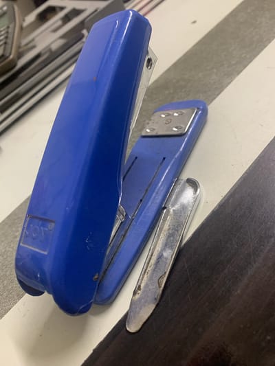 The image depicts a close-up view of a blue stapler and a silver staple remover on a striped surface, likely a desk. The stapler is open, revealing its metallic components. The staple remover is positioned alongside the stapler, with its curved edge facing upwards. The background shows a blurry, out-of-focus area that appears to be a collection of metallic objects, possibly computer components or office equipment. The lighting suggests an indoor setting, with some light reflecting off the metallic surfaces of the objects. The scene is devoid of any individuals or specific landmarks that would indicate its location within Angeles City, Philippines.
