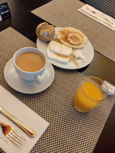 The image depicts a breakfast setting on a dark brown rectangular table with two light brown and dark brown woven placemats, indicative of an indoor dining environment, likely within a hotel or guesthouse in Porto, Portugal.
Centrally positioned is a white ceramic plate containing a single crusty brown bread roll, three small golden-brown pancakes or fritters, and several rectangular slices of white cheese. All items on the plate, specifically the pancakes and cheese, are drizzled with a golden-brown liquid, consistent with honey. To the immediate left of the plate, a white ceramic cup filled with a light brown beverage, likely coffee with milk, rests on a matching saucer.
To the right of the plate, a clear glass with a textured base contains a vibrant orange liquid, consistent with orange juice. Adjacent to the juice glass, a small white packet of "PRIMOR" salted butter is partially visible, displaying Portuguese text. In the upper left corner of the table, a black stand-up card features white text, including "Menu" and "Bom dia!" (Good morning in Portuguese).
On the lower left, a silver-colored fork and a knife with a golden-toned handle are neatly arranged on a folded white napkin. Another set of a silver-colored fork and knife are visible in the upper right, also resting on a folded white napkin. No individuals are present in the frame.
