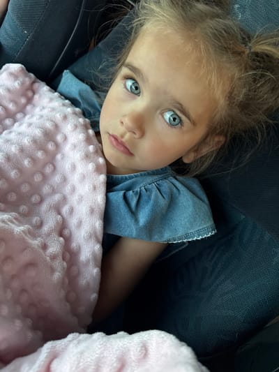 A young girl with bright blue eyes and blonde hair pulled into pigtails is looking directly at the camera. She is wrapped in a pink, fuzzy blanket with raised polka dots. She is wearing a blue denim dress with ruffled sleeves. The background appears to be dark upholstery, possibly from a car seat. There are no visible elements that suggest a specific location within the city of Étretat, France.