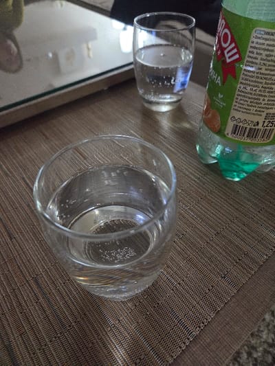 This interior scene, situated in Gevgelija, North Macedonia, depicts a tabletop arrangement. In the immediate foreground, a clear glass tumbler contains a clear liquid with visible effervescence, resting on a textured placemat woven in brown and beige tones. Adjacent to it, a 1.25L translucent plastic bottle with a prominent green label, featuring Cyrillic text (partially reading "МОЩ"), citrus fruit graphics, and a barcode, holds a pale green beverage. Its base is also green. Further back and slightly to the left, a second clear glass, also filled with a clear, carbonated liquid, is positioned on a reflective surface, identified as a glass tabletop, which shows indistinct reflections of the surrounding environment. A lighter, horizontal surface is visible in the distant background. No individuals or active interactions are present in the image.