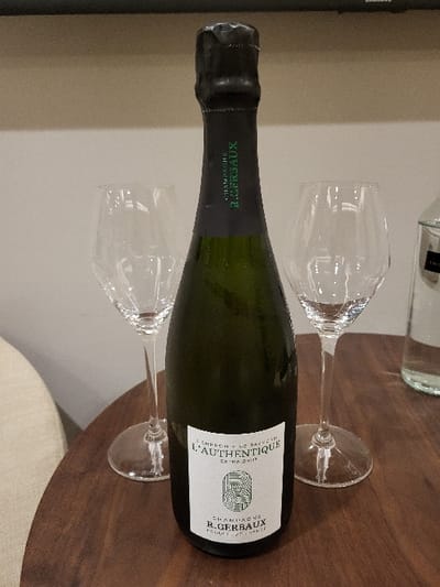A champagne bottle, labeled "Champagne R. Gerbaux L'Authentique Extra Brut," lies horizontally on a wooden table. Two empty champagne flutes flank the bottle. Another bottle, possibly of water, is partially visible in the background. The scene is set against a neutral wall, and a cushion peeks into the frame on the bottom right.