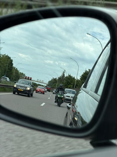 This is a photograph taken from the perspective of a driver's side mirror, showing traffic on a highway in Mondercange, Luxembourg. The mirror reflects a cloudy sky with streaks of sunlight breaking through. On the highway itself, a green Kawasaki motorcycle with a rider in a black helmet and racing suit is in motion. Behind the motorcycle, a silver Volkswagen hatchback is visible. Further ahead, a black Nissan Qashqai is in the left lane, and a red Renault Clio is in the right lane. A red truck is also visible in the distance. The highway is flanked by trees and lampposts. The image is dated June 27, 2023, at 11:44 AM.