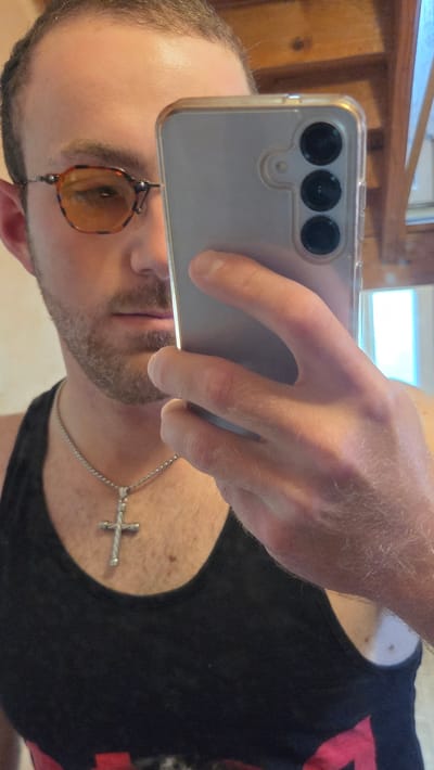 The image shows a man taking a selfie in what appears to be a home environment. He is wearing a black tank top and a silver chain with a cross pendant. He is also wearing amber-tinted sunglasses. The man has short brown hair and stubble. A cell phone, held by his hand, is visible in the foreground, with its camera array pointed towards the viewer. The background shows wooden paneling and a window, suggesting an indoor setting. The lighting is moderate, and there are no indications of time of day or weather. The overall impression is casual and personal. There is no visible text in the image.