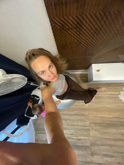A woman is sitting on a large exercise ball, her legs are bent, and her feet are flat on the floor. She is wearing a grey crop top and brown pants. Next to her, a baby in a pink onesie is reaching out. Behind the woman, there is a white fan and a dark blue surface. The floor is made of wooden planks. The ceiling has a patterned texture and appears to be angled. In the background, a white rectangular object is visible. The overall scene suggests an indoor setting, possibly a home.