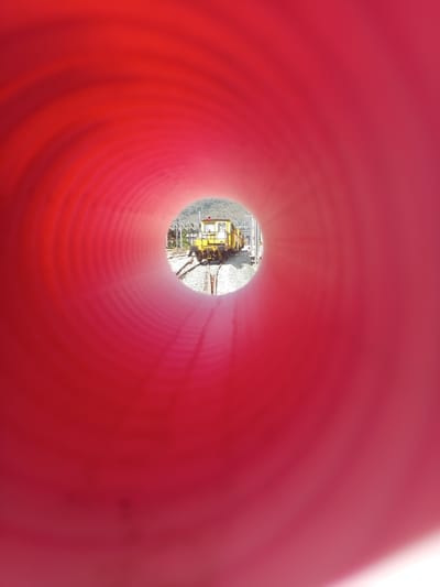 The image captures a view through a red, ribbed tunnel opening. In the distance, framed by the tunnel's aperture, a yellow train engine sits on a railway track. The train is positioned in a rural or industrial setting, with overhead power lines and a hilly, vegetated landscape visible in the background. The lighting suggests it is daytime. The tunnel opening is the dominant feature in the foreground, with its red color and textured surface creating a strong visual frame for the distant scene.