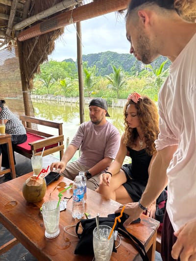 Three people are seated at a wooden table in an outdoor cafe with a thatched roof and a view of a calm body of water and lush green mountains. On the table, a whole coconut with a straw, two glasses with straws, and a bottle of water are visible. A man in a pink shirt and a baseball cap is looking down at his phone. Next to him, a woman with curly brown hair, wearing a black dress and a pink flower accessory in her hair, is looking downwards. A third person, wearing a pink shirt, is partially visible on the right, facing away from the camera. In the background, palm trees and other foliage line the water's edge. The scene suggests a relaxed outdoor dining or drinking experience.