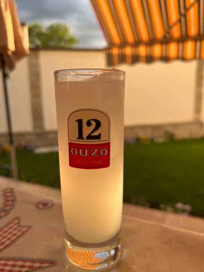 A horizontal shot features a glass of chilled Ouzo 12, sitting on a patterned tablecloth, presumably outdoors. The background is somewhat blurred, showing greenery and parts of a building, alongside an orange and white striped awning. The location, Markovo in Bulgaria, is not explicitly visible within the image.