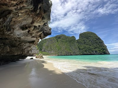 This image depicts a tropical beach scene with dramatic limestone cliffs on the left and a lush green mountainous island in the background, likely located in the vicinity of Ao Nang, Thailand. The foreground features a pristine white sand beach with gentle waves lapping at the shore, creating a foamy white border. The water transitions from a clear turquoise near the beach to a deeper blue further out. Several people are visible in the distance on the beach, appearing as small figures against the vast landscape. The sky is a vibrant blue with scattered white clouds, suggesting a sunny day. The overall environment is serene and picturesque, characteristic of popular Thai island destinations.