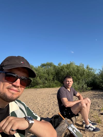 This is a selfie-style horizontal photograph taken outdoors on a sunny day. Two men are visible. The foreground is dominated by the face of a man wearing a black baseball cap and sunglasses. He is smiling and looking towards the camera. He is wearing a black t-shirt underneath a white and green striped sweater. A silver watch with a dark face is on his left wrist. A silver ring with a dark stone is on his right ring finger.
In the background, another man is lying on sandy ground, propped up on his elbows. He is wearing a dark grey t-shirt and black shorts with a pattern on them. He is looking towards the camera with a neutral expression. He is wearing white socks and dark blue and white sneakers. To his right, a piece of metal equipment, possibly for grilling, is visible, along with a wooden handle.
Behind the men, lush green trees form a dense backdrop against a clear blue sky. The sandy ground suggests a beach or riverbank setting. The bright sunlight and clear sky indicate it is daytime, likely midday or early afternoon. The overall mood appears relaxed and casual, suggesting a leisurely outing. The location context provided is Arkhangelsk, Russia. There is no visible text in the image.