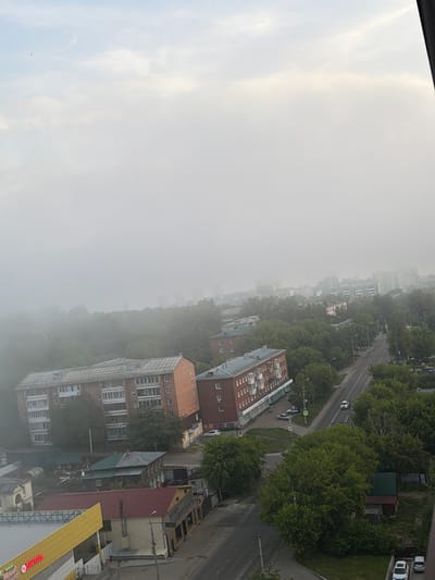 This is an aerial, medium-distance shot of a city street in Irkutsk, Russia, obscured by heavy fog or mist. The foreground features the top of a yellow and white building with red lettering on its side. Further back, a two-story building with a green roof is visible, next to other structures and trees. A road runs diagonally through the midground, with a few cars visible on it. Two large, brick apartment buildings with multiple stories and visible windows are prominent in the left-midground. Beyond these buildings, the cityscape recedes into the fog, with taller structures and trees creating a hazy silhouette. The sky above is overcast and bright, with some clouds faintly visible. There are no discernible people or animals in the image. The overall impression is one of a quiet, possibly early morning scene in a residential area.