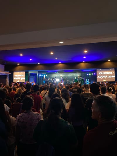 The image shows a large crowd of people gathered indoors, likely at a concert or event in Valencia. A stage with performers and lighting is visible in the distance. A screen displaying "Valencia Adora 2026" suggests the event is a religious or worship gathering. The audience members are facing the stage, with some holding up their phones to record or photograph the event. Blue lights illuminate the ceiling above the crowd.