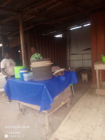 The scene depicts a semi-enclosed commercial or service area in Nsugbe, Nigeria. The structure features corrugated metal walls, some appearing brownish-red, and an exposed wooden beam roof framework. In the foreground, a wooden table is covered with a bright blue tablecloth. Positioned on this table are a lime green cooler, a blue and white lidded cylindrical container, a clear rectangular plastic container, and a large, dark-colored insulated cooler with a lid.

Two adult females are visible. On the left, a woman wearing a light-colored short-sleeved top with a dark emblem stands beside the table. In the center-right background, behind the table, another woman wearing a green and black patterned top is present. A third individual, partially obscured by the blue tablecloth and the large cooler, can be seen sitting or stooping behind the table, wearing brown and white striped clothing.

In the background, a light-colored wooden bench is situated against a wall. To the far right, a simple wooden stand holds a small, lime-green plastic bucket. The ground surface is light-colored concrete or pavement. Natural light illuminates the area. The image carries a timestamp of 2026/03/06 08:42.
