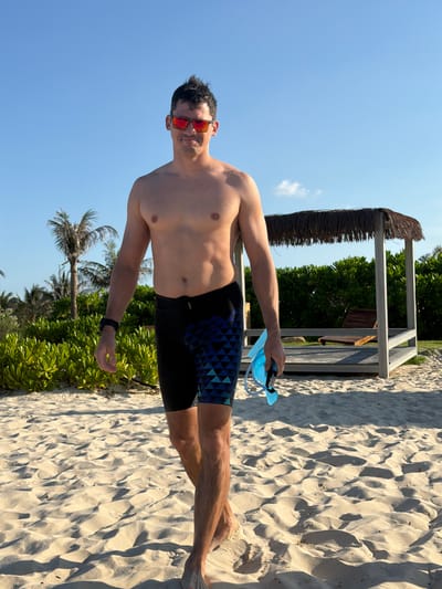 A man walks across a sandy beach on a sunny day. He is shirtless, wearing dark swim trunks with a blue geometric pattern, red-tinted sunglasses, and a black wristwatch. He is holding a blue snorkel and mask in his right hand. In the background, palm trees and lush green bushes are visible, along with a thatched-roof gazebo and a wooden lounge chair. The sand is marked with footprints and ripples from the wind.