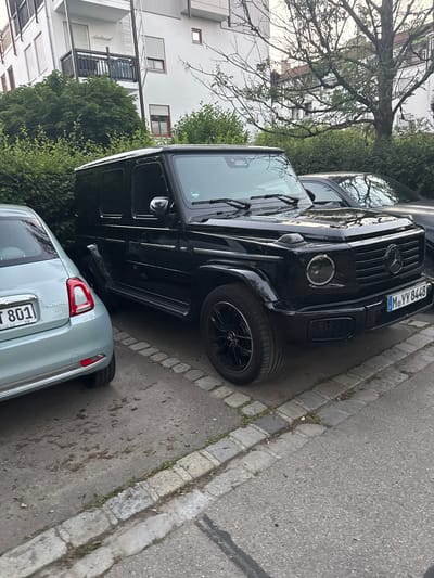 This is an outdoor, eye-level, medium shot image taken on a street in Höhenkirchen-Siegertsbrunn, Germany. The scene depicts two parked cars and a residential building. The primary subject is a black Mercedes-Benz G-Class, parked on the right side of the frame. It is facing forward, with its front right tire and fender clearly visible. The car has a German license plate that reads "M YY 8448". To the left of the G-Class, a light blue Fiat 500 is parked, facing to the right. Its rear bumper, taillight, and rear license plate, which reads "T 801", are visible. A portion of the side of the building is visible in the background, with windows and a balcony. The ground is paved with asphalt and cobblestones. There are trees and bushes surrounding the area. The lighting suggests it is daytime, possibly late afternoon or early evening, with soft lighting and some shadows. There are no people visible in the frame.