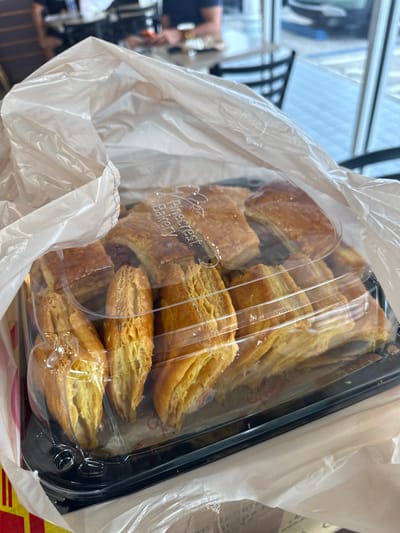 The image shows a clear plastic container filled with pastries from Pinecrest Bakery, inside a white plastic bag. The container has a black rim. The pastries appear to be flaky and golden brown. The background is blurred, showing what seems to be a window with a view of an interior space, possibly a store or cafe. Given the presence of Pinecrest Bakery, it can be inferred that the location is likely in Miami Beach, United States of America, where this bakery has multiple locations.