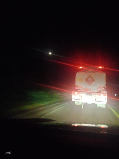 A tanker truck is driving on a road at night. The truck is white with a red and orange hazard symbol on the back. It has red taillights and flashing orange lights on top. The road appears to be a highway, with a yellow line marking the lane. There is some greenery on the side of the road. The moon is visible in the sky. The image was taken from inside another vehicle. The location is Seunebobaru, Indonesia.