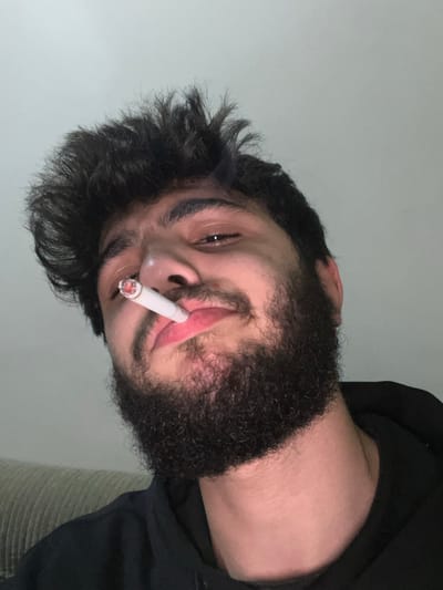 A man with dark, curly hair and a thick, dark beard holds a lit cigarette in his mouth. He looks slightly upwards, toward the camera. His skin is fair, and his expression is relaxed. He appears to be wearing a dark-colored top or hoodie. The background is a plain, light-colored wall. The image has a slight tilt to it.