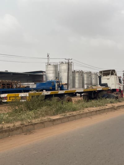 The image captures an industrial roadside scene in Ota, Nigeria, under an overcast sky. In the background, a complex features a row of approximately six large, cylindrical, silver-colored industrial storage tanks with visible catwalks, alongside a dark-roofed industrial building and stacks of wooden pallets. A metallic communication tower stands behind the tanks. Overhead, numerous power lines stretch between utility poles. In the midground, two flatbed trucks are parked on a reddish-brown dirt shoulder. The truck on the left has a blue cab and a flatbed trailer with a prominent yellow and white striped rear bumper. The truck on the right features a white and red cab, appearing as a tractor unit. The dirt shoulder is overgrown with green and brown weeds and grass. The foreground consists of a concrete curb delineating the shoulder from a dark asphalt road. No individuals are present.