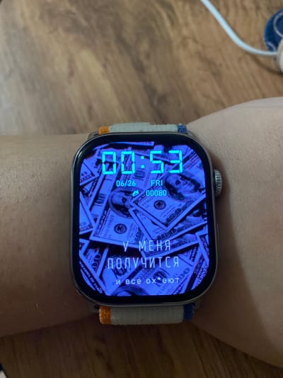 The image shows a close-up of a smartwatch worn on a person's wrist. The watch face displays the time as 00:53, with the date 06/26 and the day of the week FRI. The background of the watch face is an image of U.S. dollar bills. Below the time and date, there are Russian words that translate to "I will succeed" and "and all will be". The watch has a grey metallic casing and a fabric band with blue and orange stripes. The background of the image is a wooden surface, possibly a table or floor. There is no indication of the location being Pokhvistnevo, Russia.