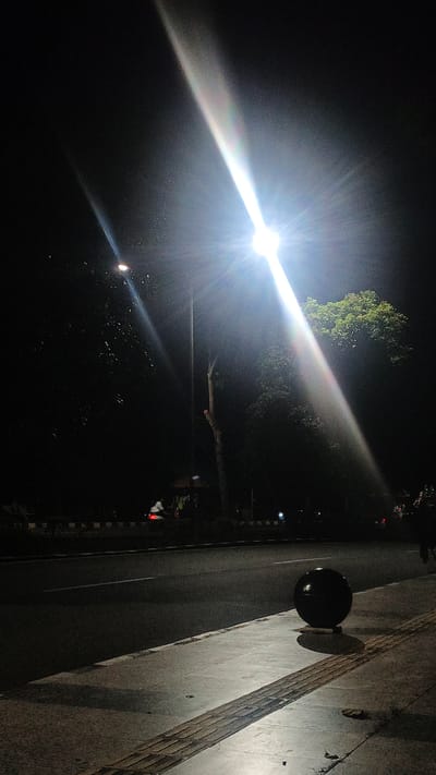 This media file depicts a nighttime scene on a street in Sodong, Indonesia. The primary subject is a bright street lamp casting a strong, angled beam of light across the frame. The light source itself is intensely bright, creating a starburst effect and a prominent lens flare that splits into a spectrum of colors, suggesting the presence of moisture in the air, perhaps from recent rain or humidity.
The scene includes a road with a white line marking the center, suggesting it's a public thoroughfare. On the right side of the road, a sidewalk is visible, constructed with textured tactile paving blocks, typical for pedestrian areas. A large, dark, spherical object rests on the sidewalk, casting a distinct shadow. Further down the road, silhouetted trees and foliage are visible, with the dark shapes of buildings or structures in the background. A few distant lights indicate ongoing activity, and a faint silhouette of a person or a scooter with headlights on can be seen on the road in the middle distance, suggesting some level of movement and life despite the darkness.
The time of day is clearly night, accentuated by the strong artificial light and the dark surroundings. The atmospheric conditions, indicated by the lens flare and potential haze, suggest a humid or perhaps slightly misty night. The overall mood is somewhat dramatic due to the intense light and shadow play. No visible text is present in the image. The location context of Sodong, Indonesia, places the scene in a specific geographical setting.