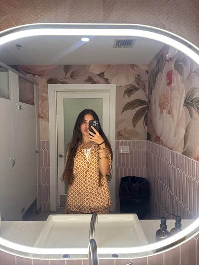 A young woman with long dark hair takes a mirror selfie in a well-lit bathroom. She is wearing a short-sleeved collared shirt with an orange and yellow geometric pattern and a white undershirt. She is also wearing several bracelets and necklaces. She is holding a black smartphone in her right hand, with which she is taking the picture. The bathroom has floral wallpaper with large pink and white flowers. There are two stalls with white doors on the left side of the image. A white sink with a silver faucet is in the foreground. There is a black trash can behind the sink and a red fire alarm on the right wall. The bathroom is located in Boca Raton, United States of America.